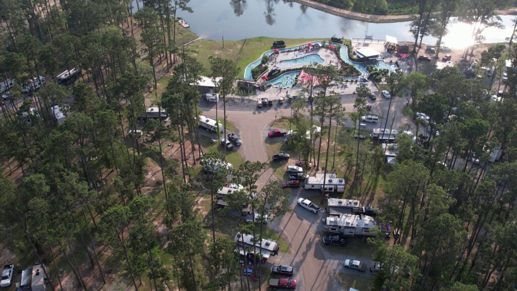 A high-angle aerial photograph captures a sprawling RV resort nestled among tall pine trees, featuring a winding water park with a lazy river and pool at Fireside RV Resort in Ponchatoula, Louisiana.