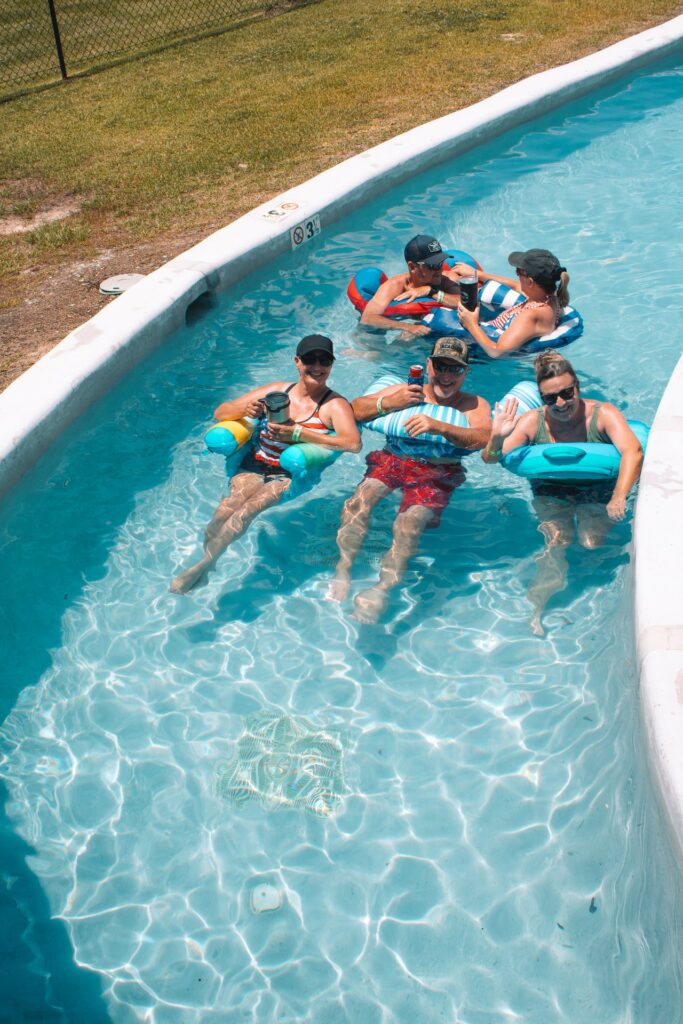 A group of five adults relaxes in a narrow, winding lazy river, floating on inflatable tubes and holding beverages. The water is clear and bright blue at Fireside RV Resort in Ponchatoula, Louisiana.