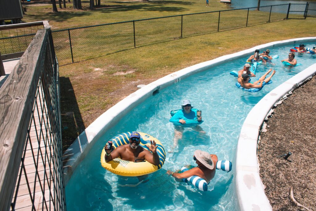 A group of people float down a winding, man-made lazy river in bright blue water on a sunny day. Several guests are using inflatable tubes and pool no at Fireside RV Resort in Ponchatoula, Louisiana.
