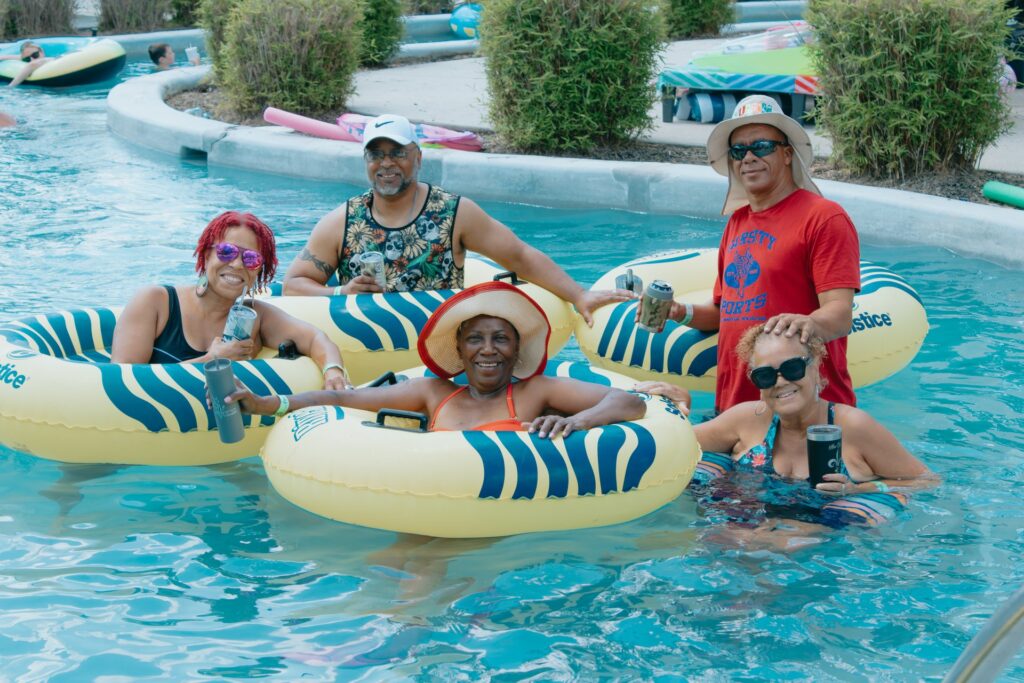 A group of five smiling adults relaxes in a lazy river, floating on yellow and blue striped inflatable tubes. They are holding beverage cans and weari at Fireside RV Resort in Ponchatoula, Louisiana.