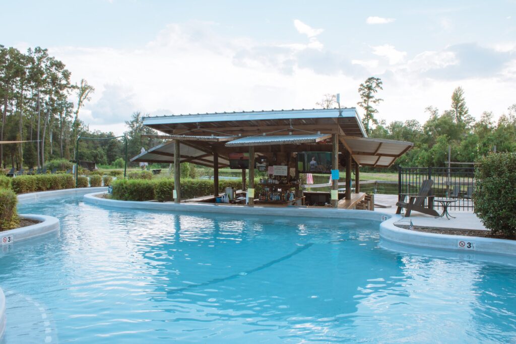 A wooden, open-air swim-up bar with a metal roof sits in the center of a winding lazy river at an outdoor resort. The bright blue water curves around at Fireside RV Resort in Ponchatoula, Louisiana.