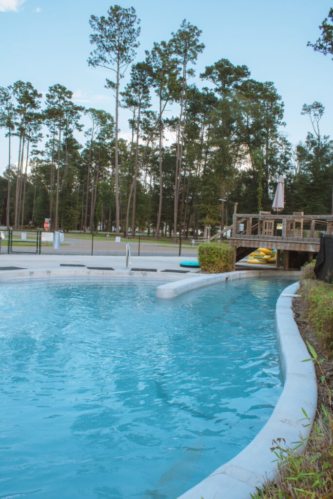 A wide shot of a resort water feature featuring a curved, light-blue lazy river channel in the foreground. In the background, a wooden bridge spans th at Fireside RV Resort in Ponchatoula, Louisiana.