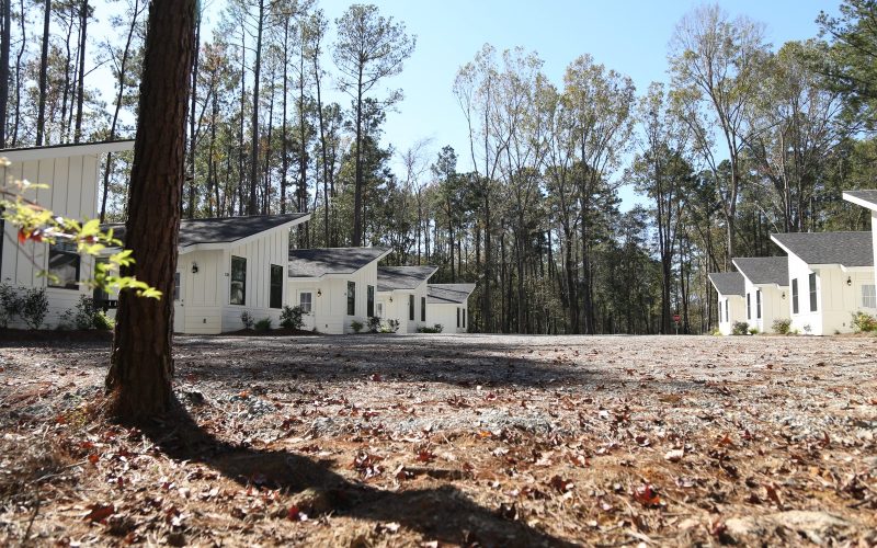 A row of modern, white cabins with dark shingled roofs sits in a clearing surrounded by tall pine trees at Fireside RV Resort in Ponchatoula, Louisiana.